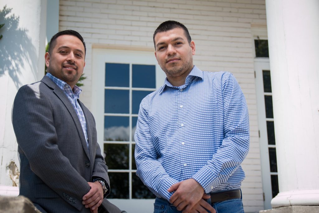 Two people with short hair pose in front of a house. They are wearing professional clothing and looking directly into the camera.