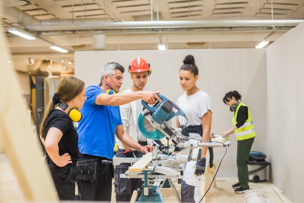 Trainees watch a carpenter demonstrate an electric saw